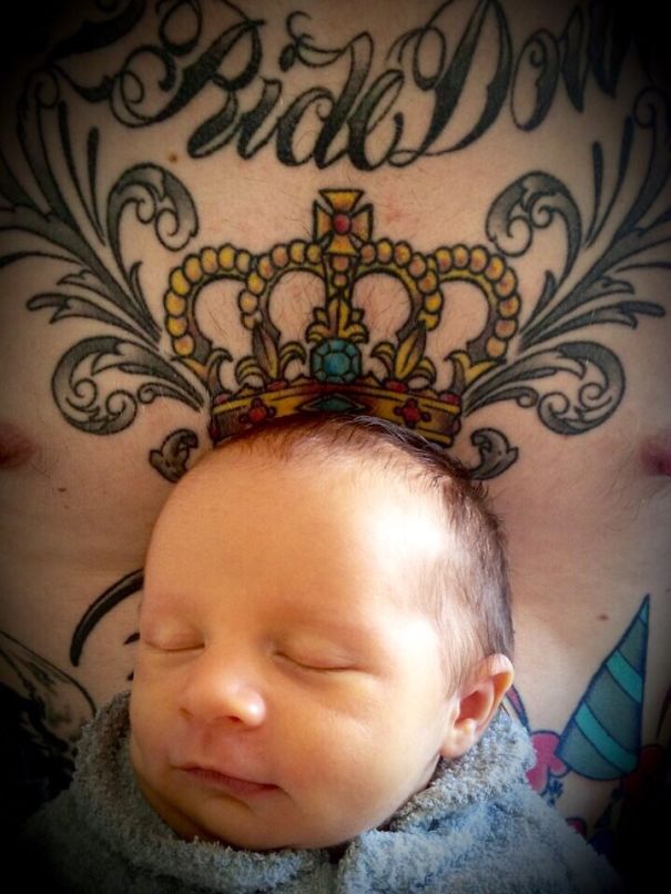 Baby sleeping peacefully against tattooed parent's chest with intricate crown tattoo.