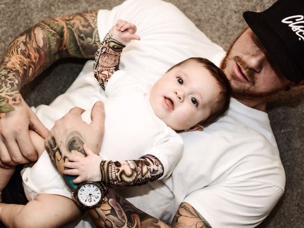 Baby with tattooed parent lying down, both wearing white, showcasing family tattoos.