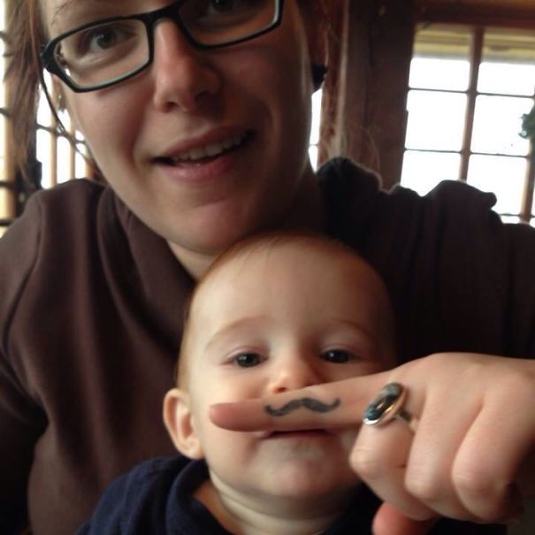 A baby posing with a tattooed parent using a finger to create a mustache effect.