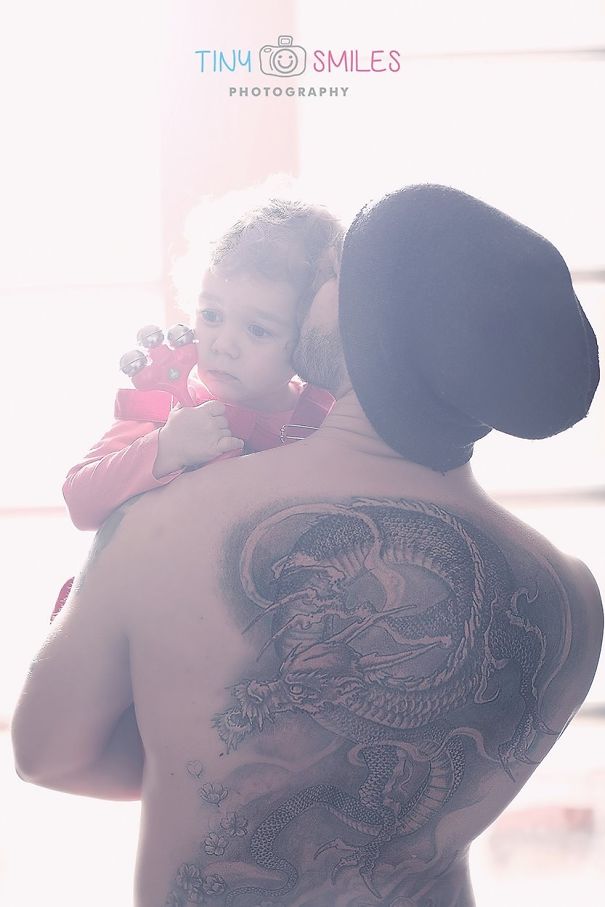 Baby held by tattooed parent with a dragon tattoo, both embracing in soft light.