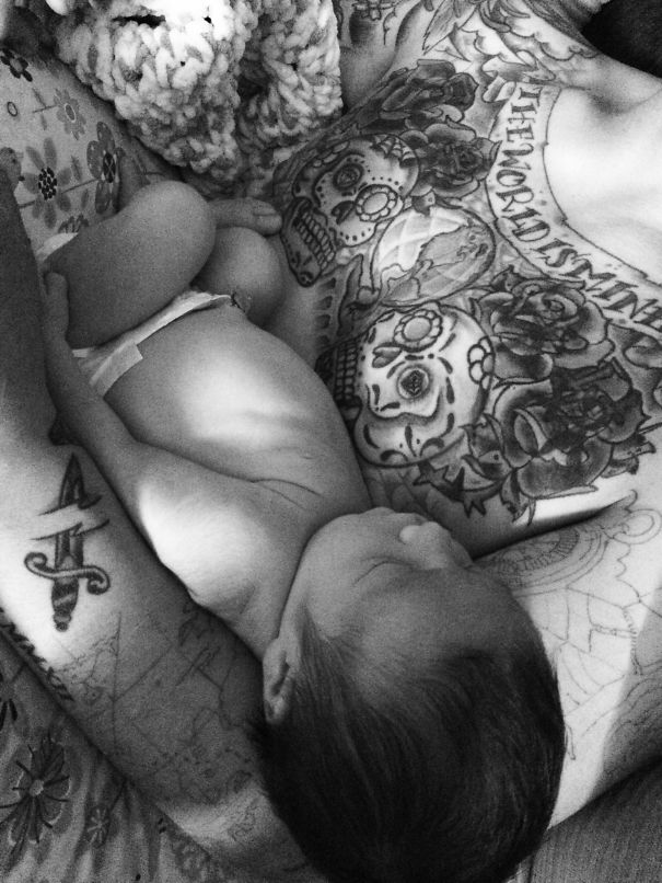 A tattooed parent cuddling with a sleeping baby, showcasing intricate chest tattoos.