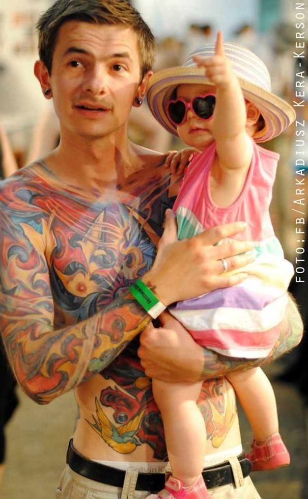Tattooed parent holding baby, both smiling at an event, showcasing beautiful bonding together.