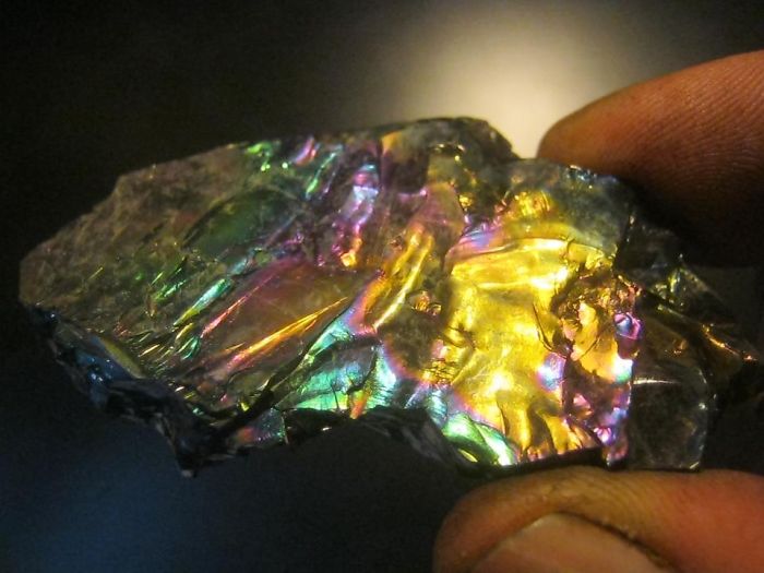 A colorful, iridescent mineral held between fingers, showcasing its extremely beautiful hues.