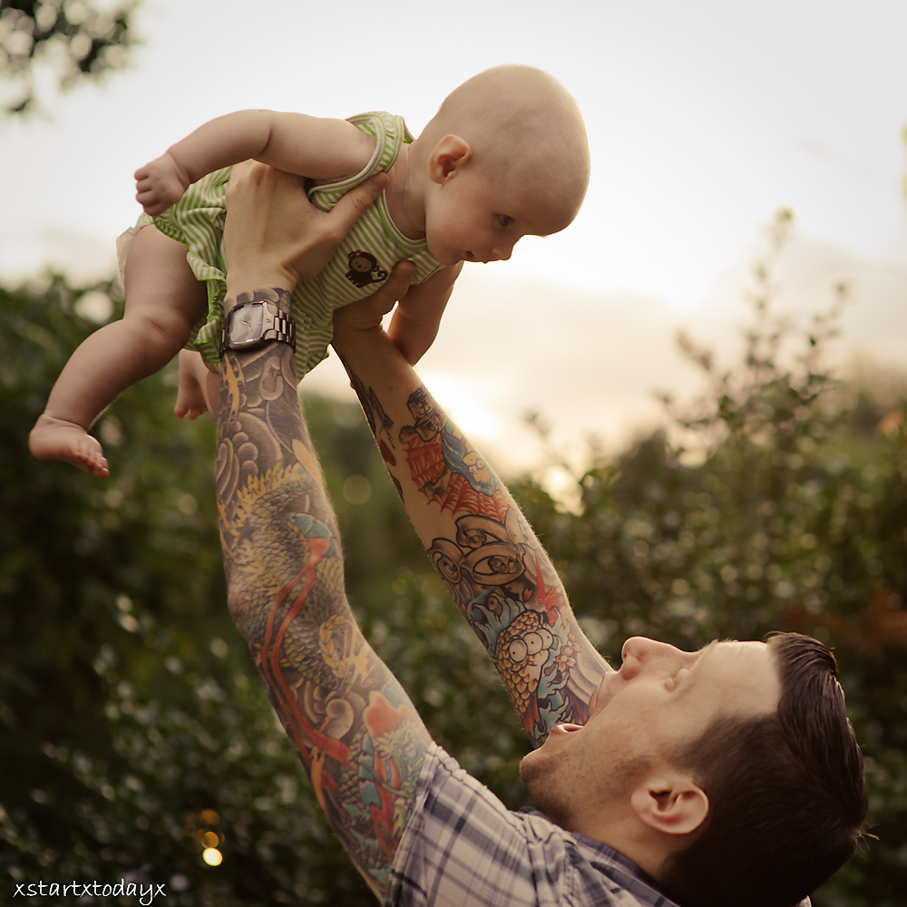Tattooed parent joyfully lifts a baby in a green outfit outdoors against a soft-focus nature background.