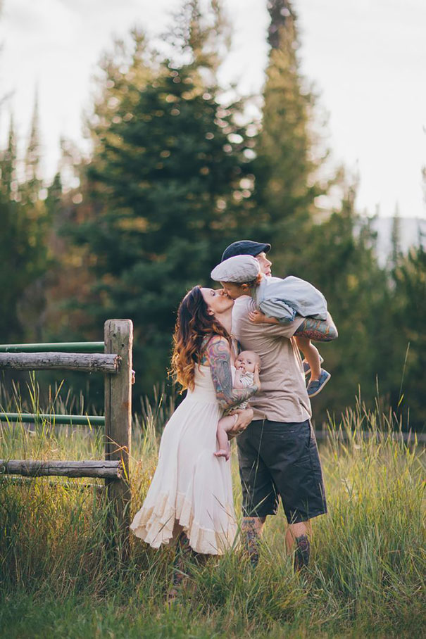 Tattooed parents share a kiss while holding their baby in a scenic outdoor setting with trees in the background.