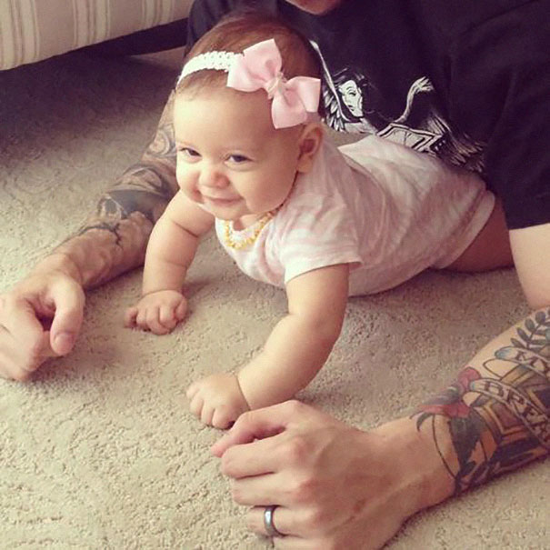 Baby crawling on carpet, next to tattooed parent, both smiling and enjoying time together.