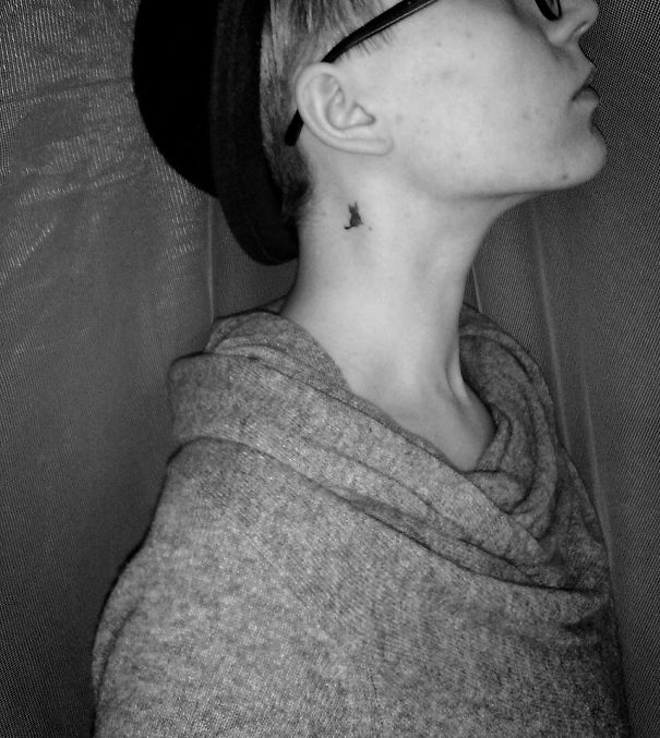 A small, creative ear tattoo on a person's neck, wearing a hat and glasses, captured in a grayscale image.