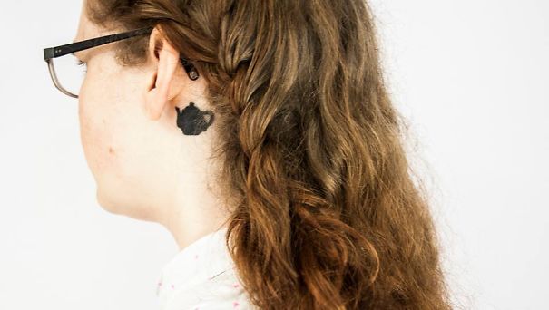 Woman with long hair showcasing a creative ear tattoo featuring a dark silhouette design behind the ear.