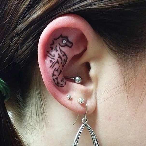 Creative ear tattoo design featuring a seahorse inside the ear, adorned with multiple piercings.