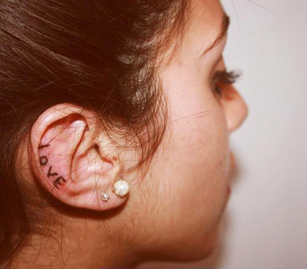 Creative ear tattoo with the word "love" inked along the ear's inner curve, next to pearl earrings.