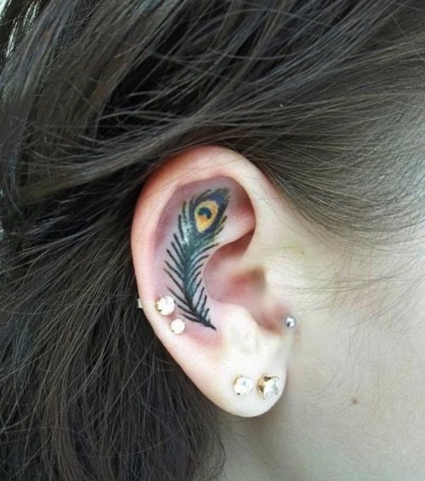 Creative ear tattoo of a peacock feather design with multiple ear piercings visible.