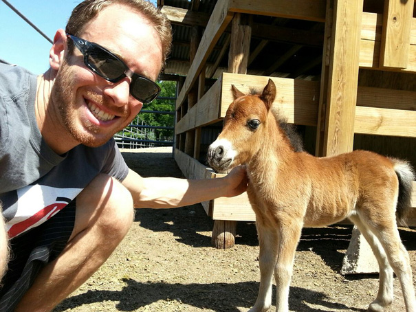 A man in sunglasses kneeling beside a cute mini horse outside a wooden barn.