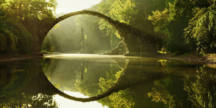 Mystical stone bridge forming a perfect circle over calm water surrounded by lush green forest in soft sunlight.