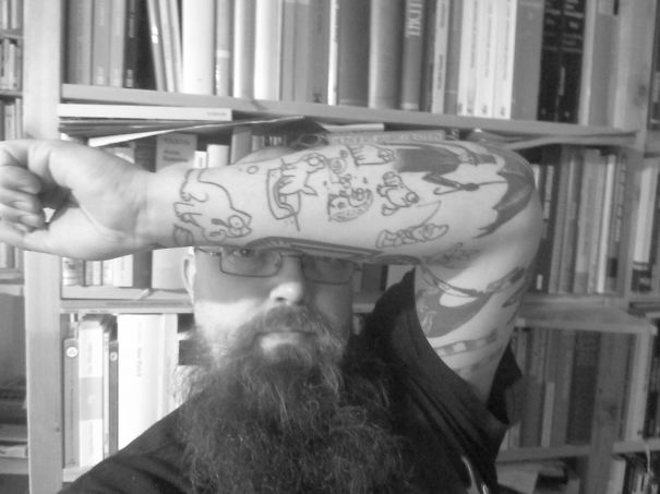 Man showcasing simple cat tattoos in line art style on his arm, in front of a bookshelf.