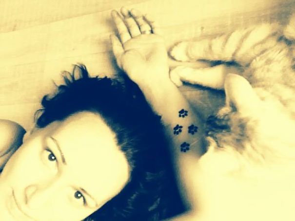 Woman with a simple cat tattoo, featuring paw prints, lying beside a cat.