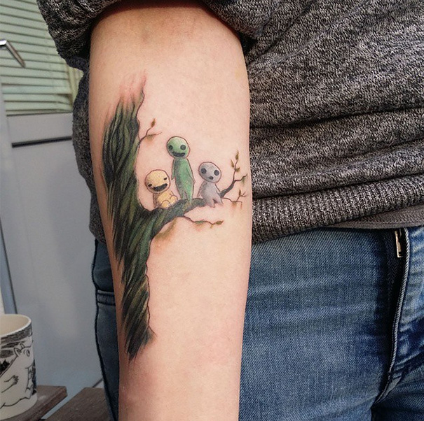 Studio Ghibli tattoo featuring Kodama spirits from Miyazaki films on a person's forearm, with colorful tree and branches.