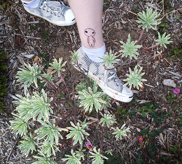 Tattoo of a Studio Ghibli Kodama on a leg wearing striped sneakers, surrounded by plants.