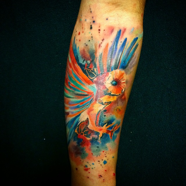 Colorful watercolor tattoo of a bird by Czech artist Ondrash, showcasing vibrant blue and orange hues on an arm.