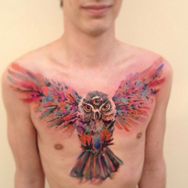 Watercolor tattoo of a vibrant owl with spread wings on a man's chest, showcasing Czech artist Ondrash's artistic style.