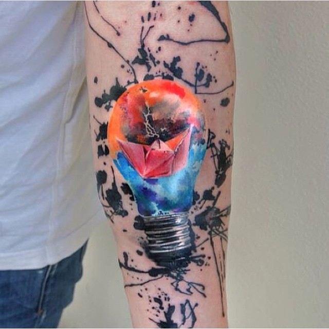 Watercolor tattoo of a lightbulb with a paper boat, featuring vibrant colors on an arm.