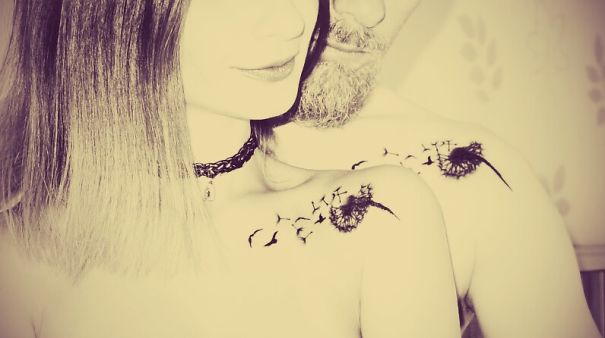 Couple with matching wedding tattoos on their shoulders, featuring dandelions and birds, embracing closely.