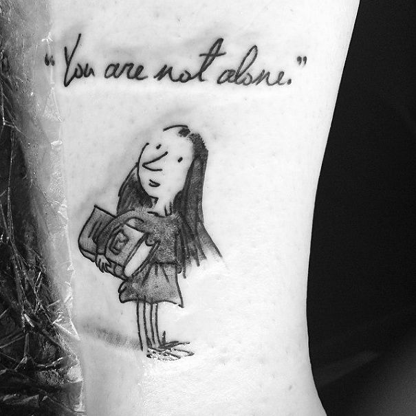 Tattoo of a girl holding a book with the quote "You are not alone" above her, inspired by literature.