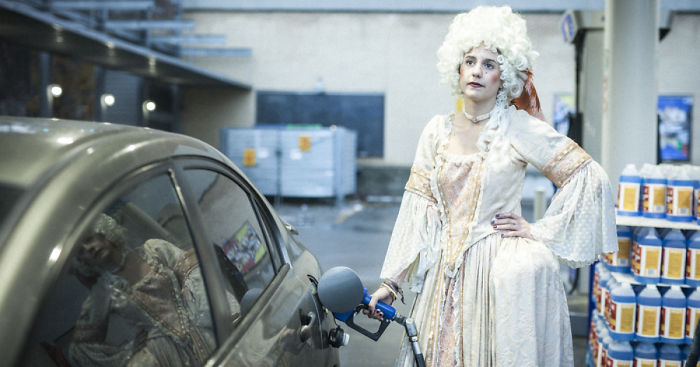 A woman in 18th century fashion with a powdered wig, filling a modern car with fuel at a gas station.