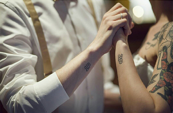 Couple with matching wedding tattoos on wrists instead of rings, holding hands in an intimate setting.