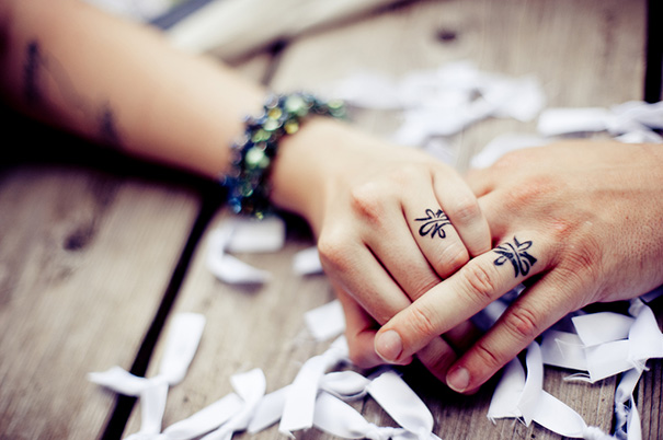 Couple's hands with matching wedding tattoos instead of rings, surrounded by paper confetti on a wooden table.