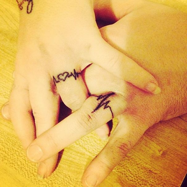 Couple's hands with matching wedding tattoos, featuring heart and heartbeat designs on their ring fingers.