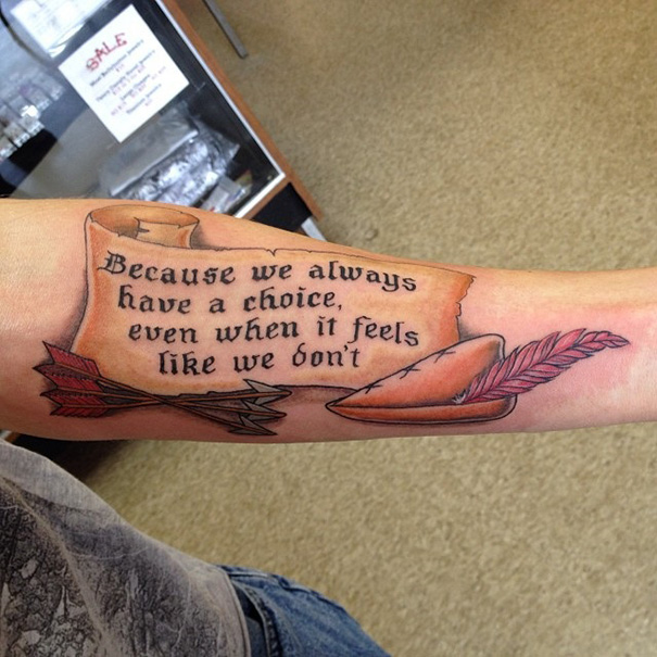 Tattoo of a parchment with a quote, arrows, and quill inspired by books on a person's forearm.
