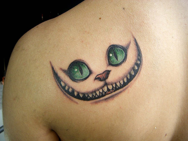 Tattoo of a Cheshire Cat on a shoulder, inspired by books, featuring vibrant green eyes and a wide grin.