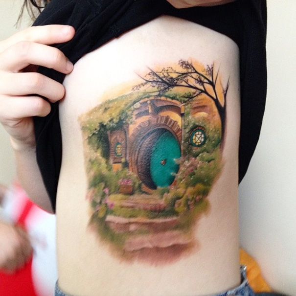 Tattoo of a hobbit house on skin, inspired by a beloved book, with detailed greenery and round door.