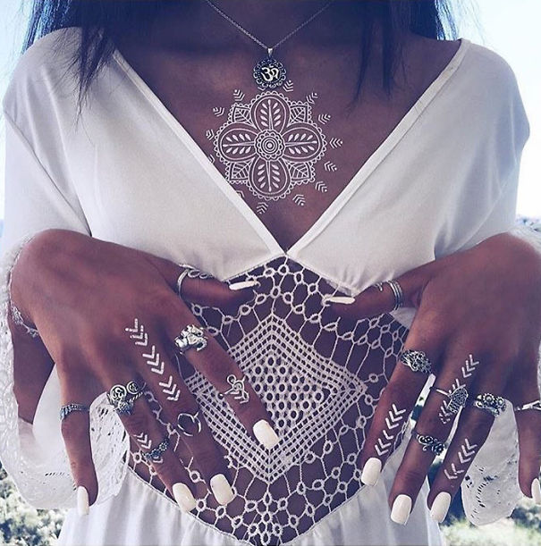 White henna-inspired tattoo on chest and hands, resembling elegant lace, with silver rings and white nails.