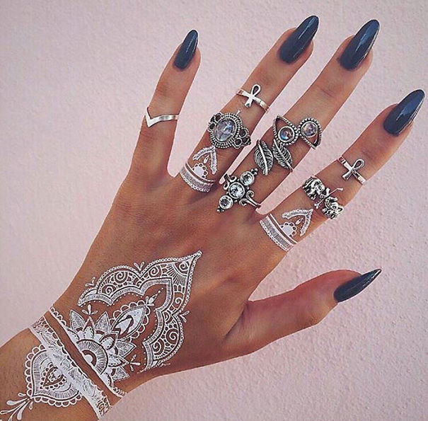 White henna-inspired tattoos resembling elegant lace designs on a hand adorned with rings.