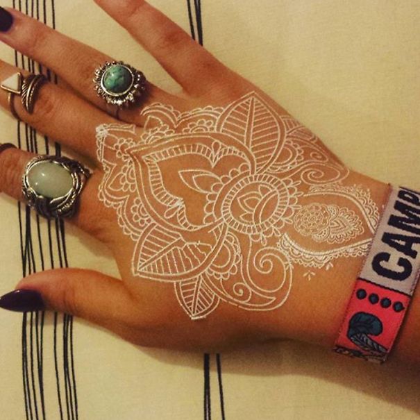 White henna-inspired tattoo on hand, featuring intricate lace-like floral designs, complemented by turquoise rings.
