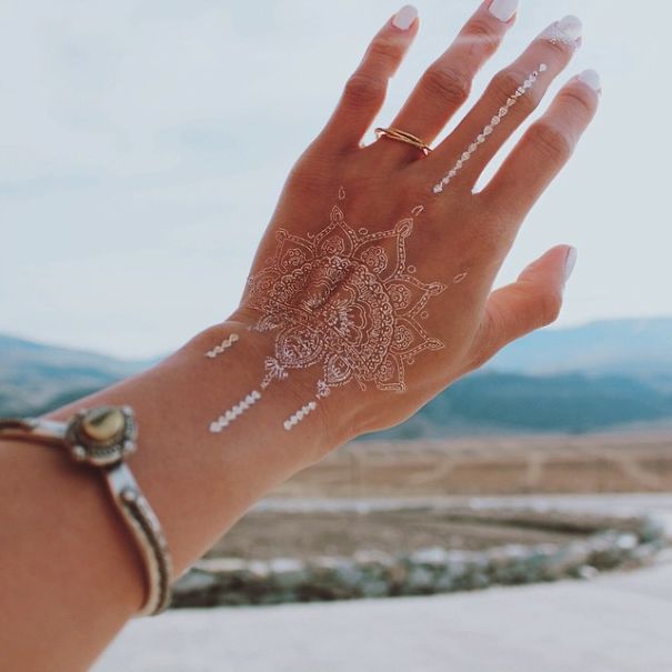 White henna-inspired tattoo on hand, resembling elegant lace, with scenic background.