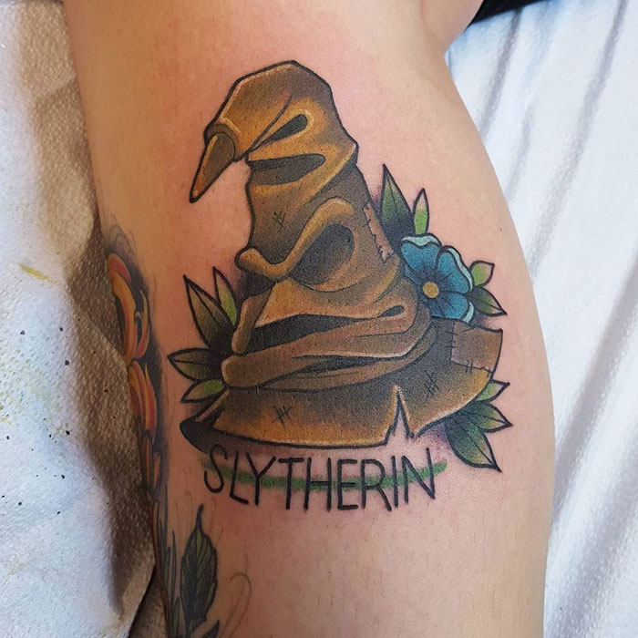 Harry Potter tattoo of the Sorting Hat with "Slytherin" text and floral design on skin.