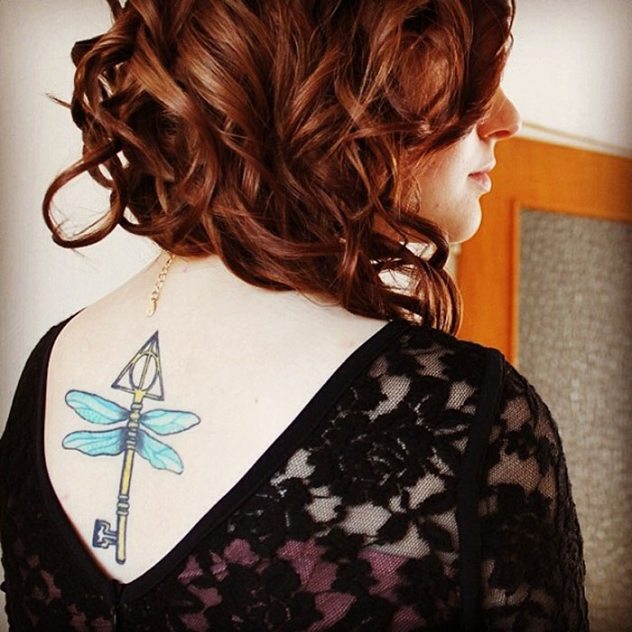 Woman with curly hair showing a Harry Potter tattoo of a winged key on her upper back, wearing a lace top.