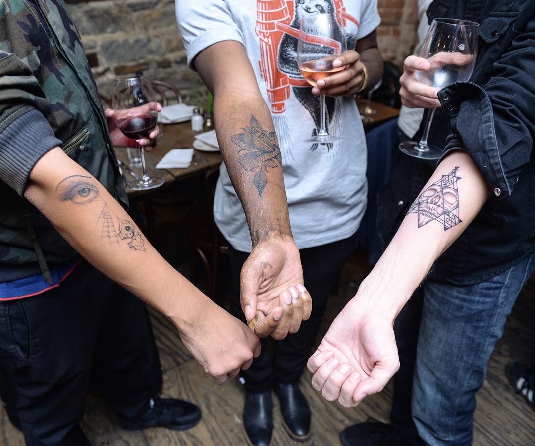 Three people show off surprise tattoos from famous artist, holding wine glasses in a casual setting. Three people show off surprise tattoos from famous artist, holding wine glasses in a casual setting.