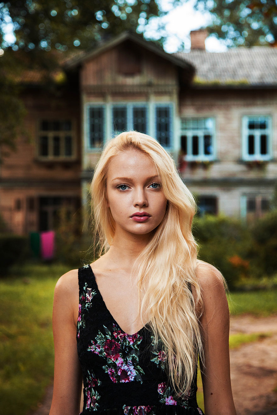 Romanian photographer captures diverse beauty, featuring a woman with long blonde hair in front of a rustic house.