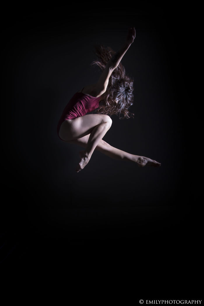 Ballet dancer in a red leotard performing a mid-air leap against a black background, showcasing the art of ballet.