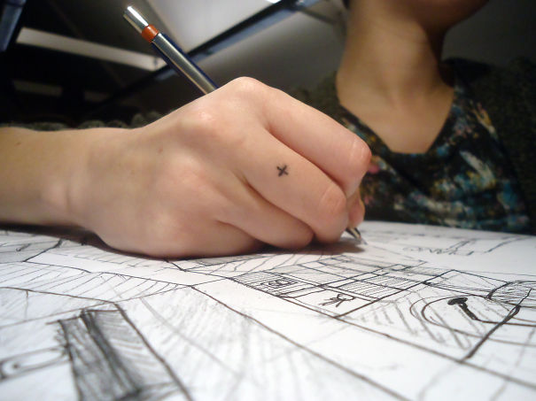 A small minimalist tattoo of an "X" on a person's hand while sketching with a pencil.