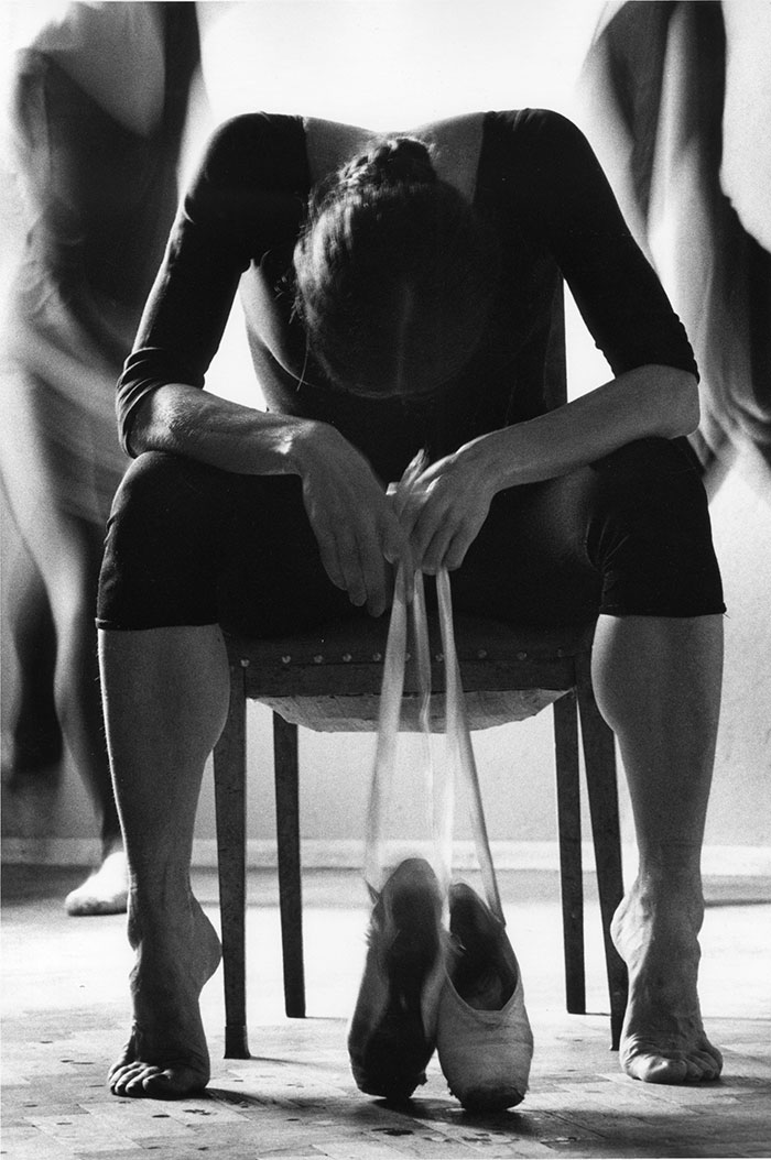 Ballet dancer resting on a chair, holding pointe shoes, capturing the cost of applause and the art's dedication.