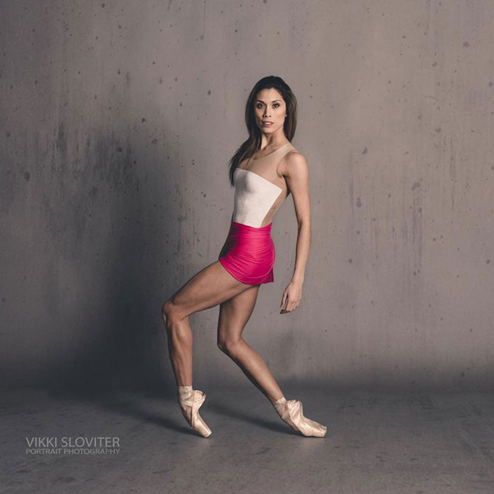 Ballet dancer in a pink skirt gracefully posing en pointe against a gray background.