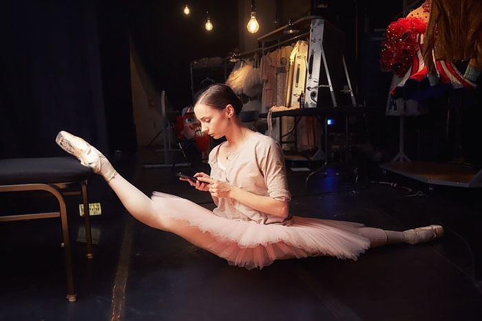 Ballet dancer in a tutu, stretching backstage while using a phone, surrounded by costumes and soft lighting.