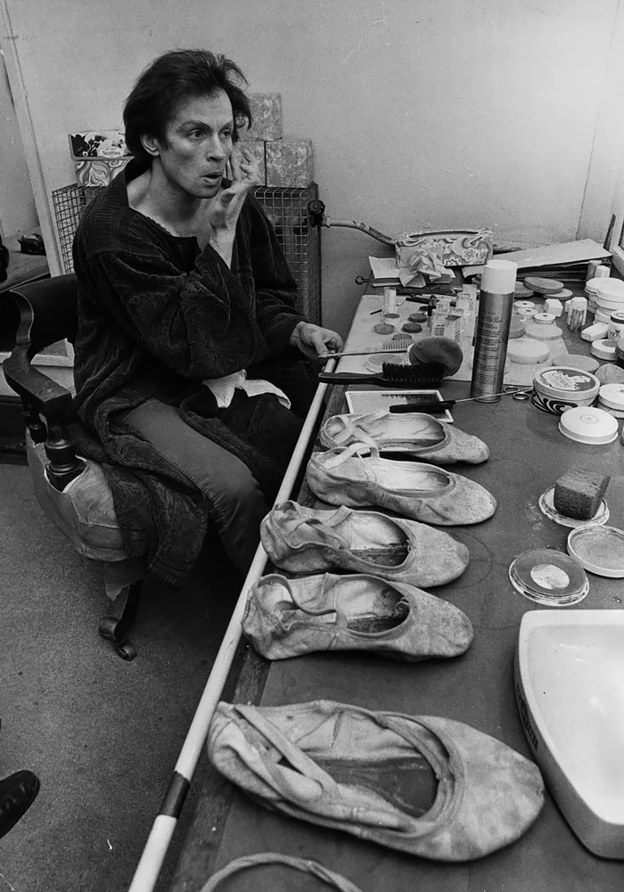 Ballet dancer backstage, applying makeup, with multiple pairs of worn ballet shoes on a dressing table.