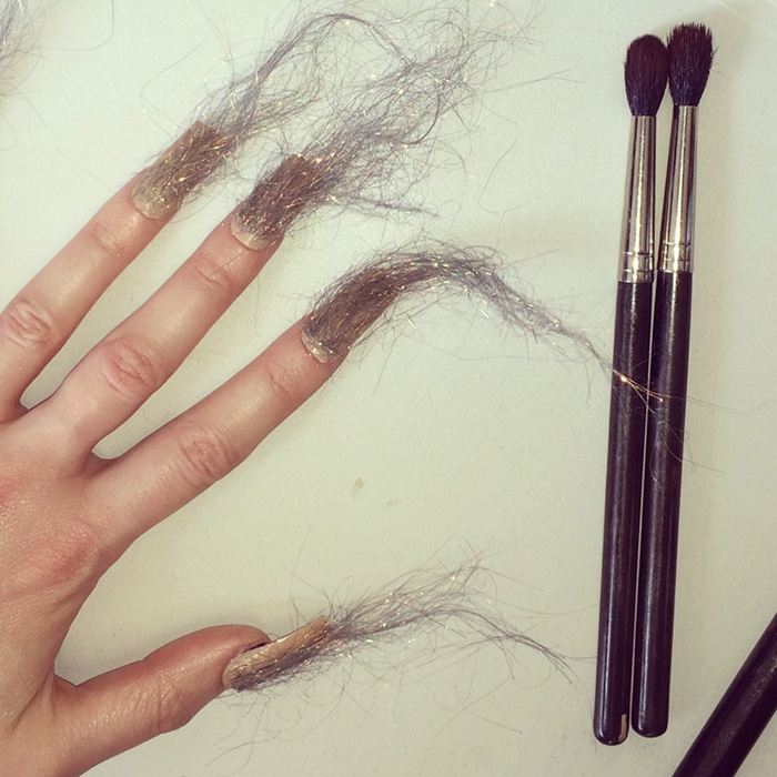 Hand with long furry nails featuring wispy hair-like fibers next to two makeup brushes on a white surface, showcasing furry nails trend.