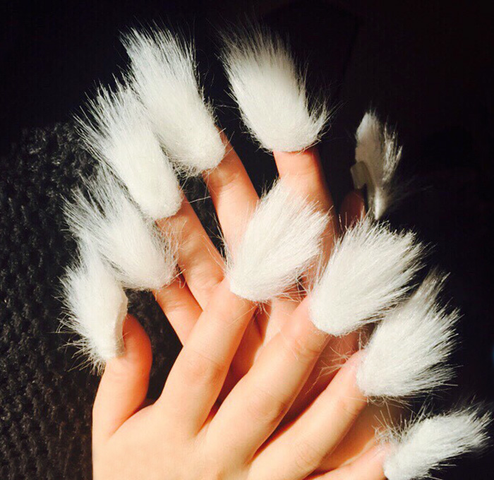 Hands displaying long white furry nails against a dark textured background, showcasing the furry nails trend.