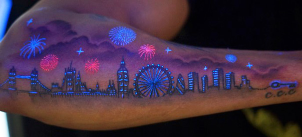 Glow in the dark tattoo of a city skyline with fireworks, visible under black light on a forearm.